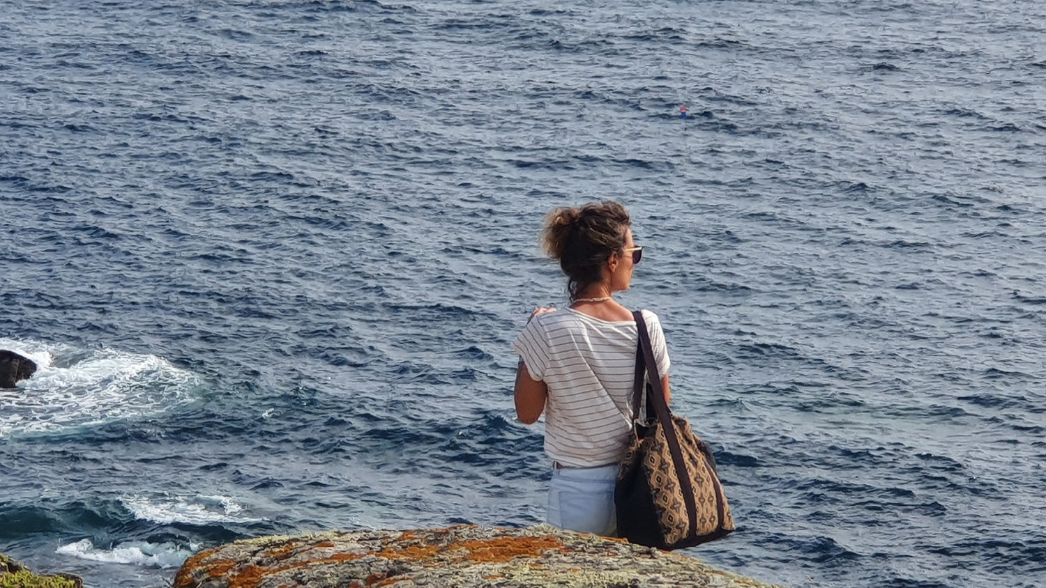Carole porte à l'épaule droite le cabas poriou devant un rochet à flanc de falaise face à la mer.